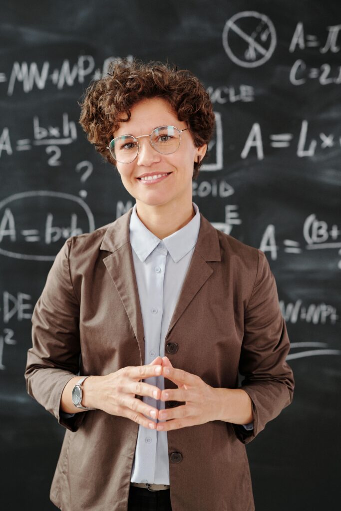 Teacher standing by a blackboard with algebra equations, smiling confidently.