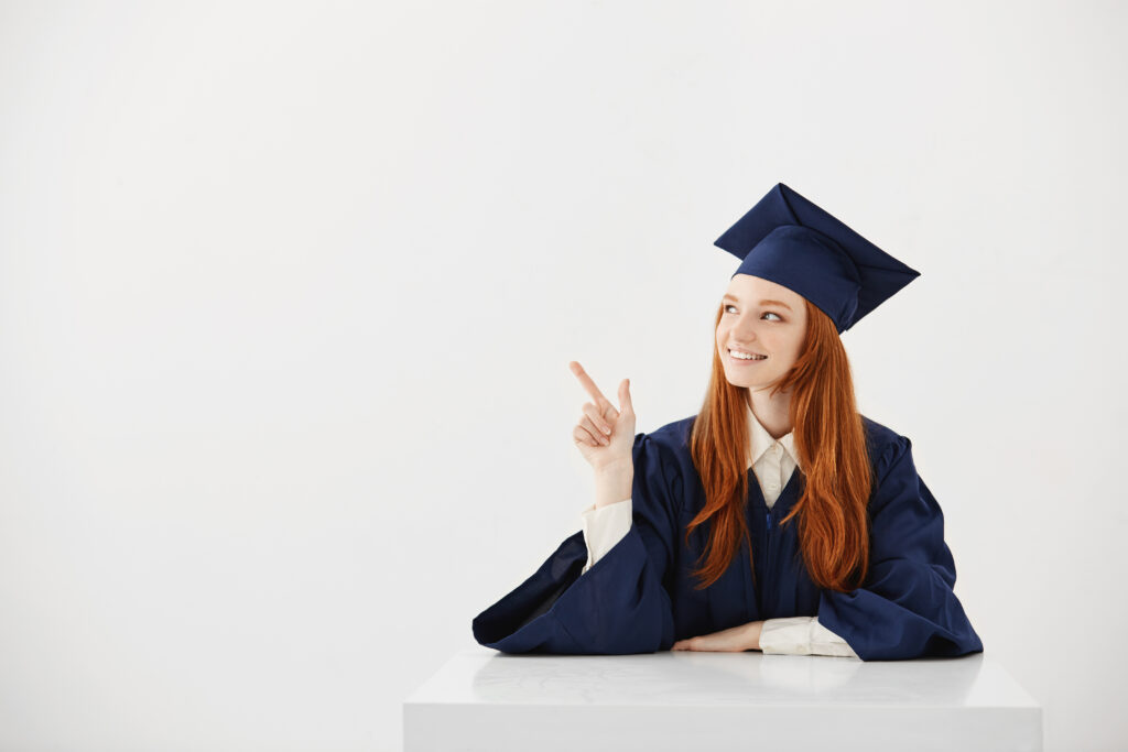 young female university graduate in academic cap sitting at table smiling pointing left. future lawyer or engineer showing an idea. white background.